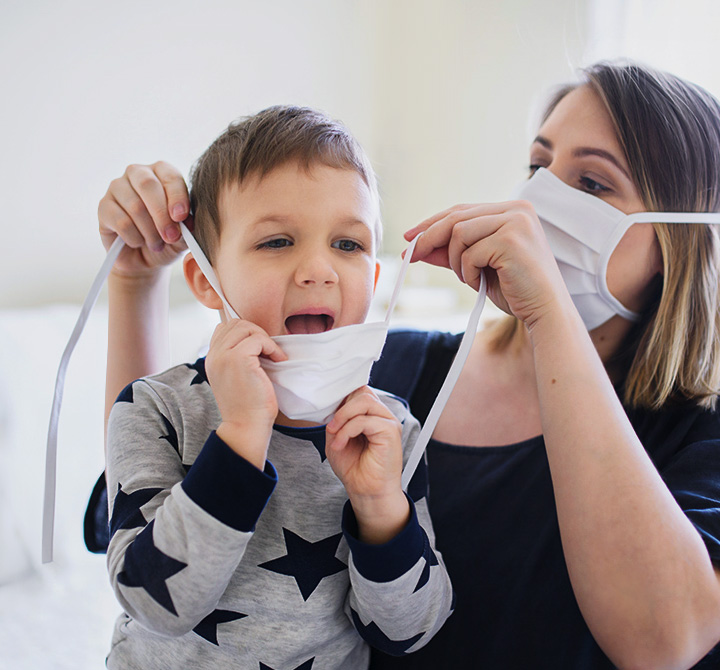 A woman is wearing a white face mask. She is helping her young son put on the same kind of mask.